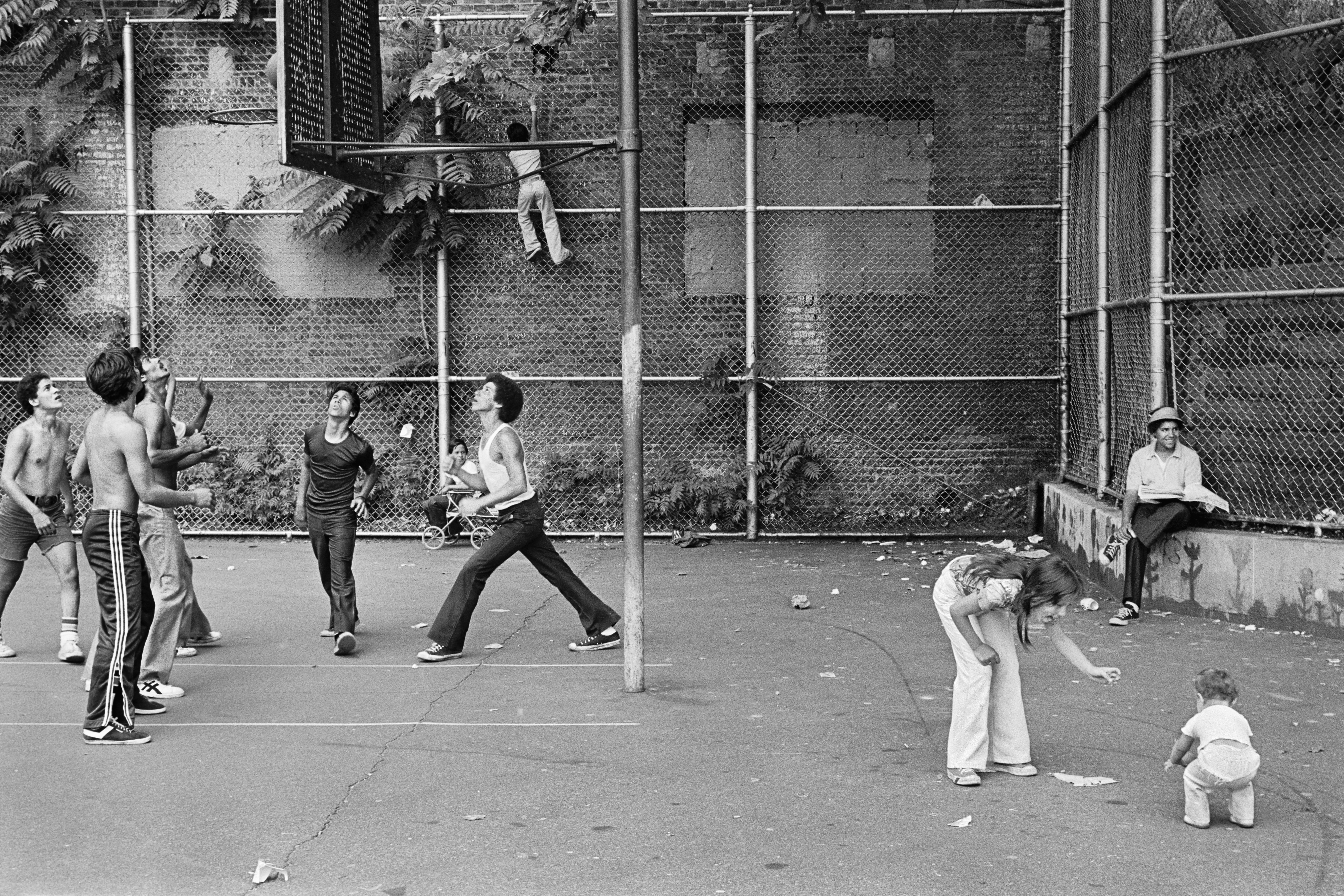 Photograph of people playing basketball in basketball court between two buildings, woman and child in front